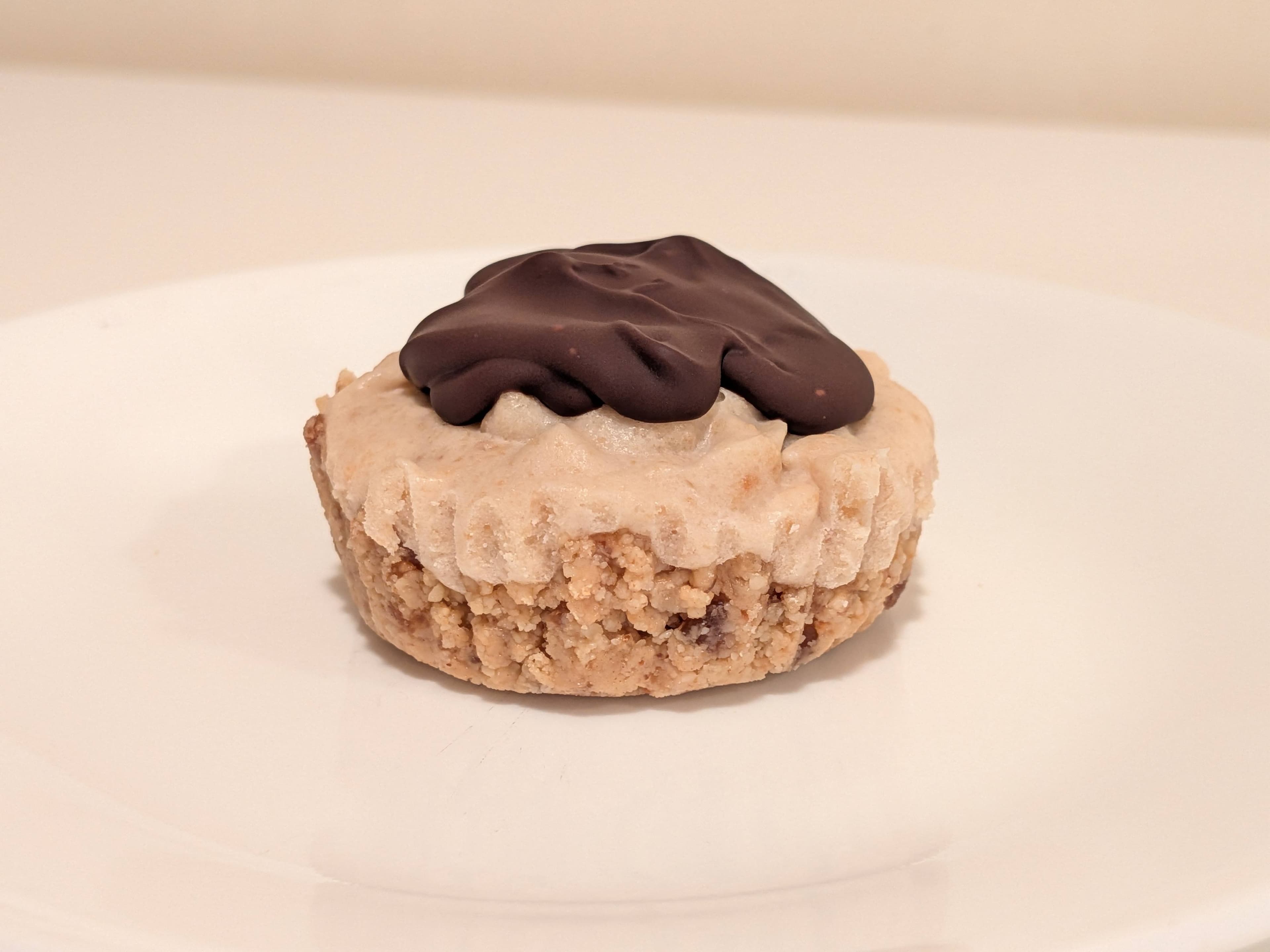 No-bake vegan peanut butter chocolate cheesecake cups in paper liners, topped with smooth glossy chocolate ganache, arranged on a silver muffin tin.